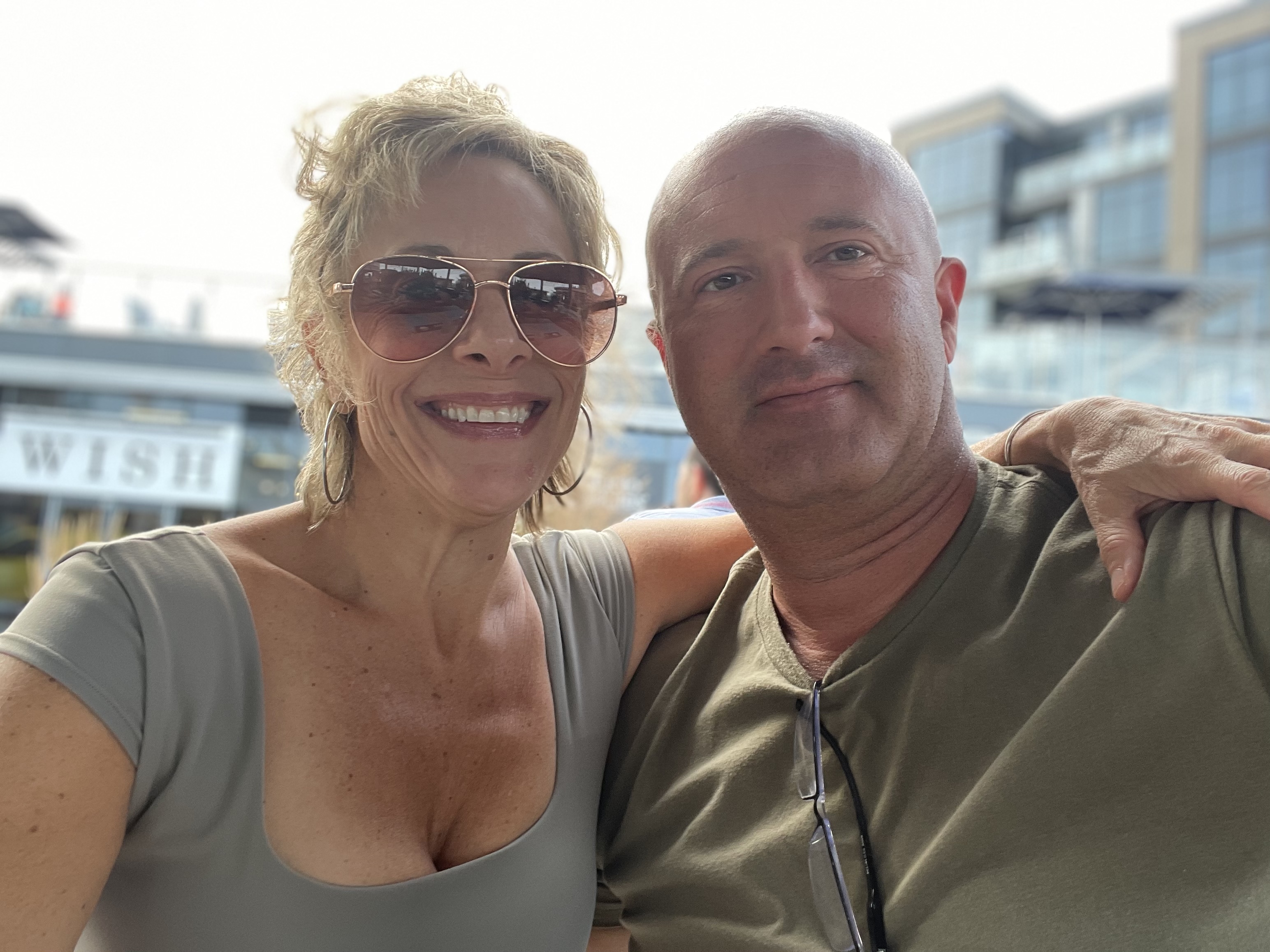 Amy and Bill smiling cheek to cheek outdoors on a sunny day, Amy in aviator sunglasses with her arm around Bill